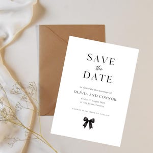 Może przedstawiać: Biała karta 'Save the Date' z czarnym tekstem, czarną ilustracją kokardy i brązową kopertą. Karta ogłasza ślub Olivii i Connora 1 sierpnia 2023 roku. Gałązka delikatnych białych kwiatów i biała tkanina w tle.
