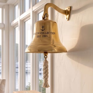 May include: A polished brass wall-mounted bell featuring an anchor icon and the text BEACON COLLECTION EST. 1995. The bell is suspended from a curved brass arm and includes a thick, braided off-white rope pull attached to the internal clapper.