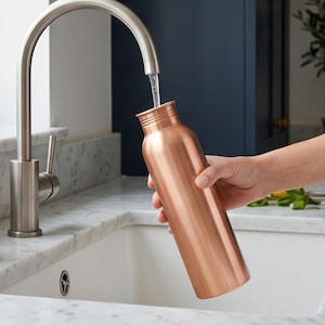 May include: A cylindrical copper water bottle with a smooth, metallic finish and a narrow neck. The bottle is held at an angle under a stainless steel kitchen faucet, with a stream of clear water flowing directly into the open top of the vessel.