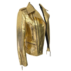May include: A gold-colored, textured leather jacket with a classic biker style. The jacket features a notched collar, multiple zippered pockets, and a front zipper closure. The sleeves have zippers at the cuffs. The jacket is a metallic gold color.