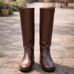 Puede incluir: Un par de botas de montar de cuero marrón, altas hasta la rodilla, con un acabado pulido. Las botas están colocadas sobre un suelo de ladrillos. Las botas de estilo ecuestre clásico están diseñadas para montar a caballo.