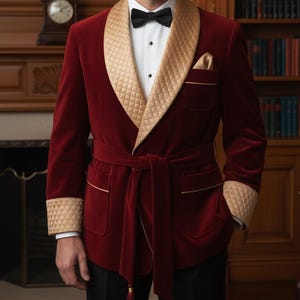 Op de afbeelding: Bordeaux fluwelen smokingjasje met een gewatteerde gouden satijnen sjaalkraag en manchetten. De jas heeft een bijpassende riem, zakdetails en een satijnen pochet. Een zwarte vlinderdas en een wit overhemd zijn zichtbaar.