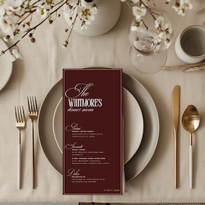 May include: A formal dinner table setting with a burgundy menu card that reads "The Whitmore's dinner menu." The menu is on a plate with gold silverware, a water glass, and a small white ceramic cup. White flowers are in the background.