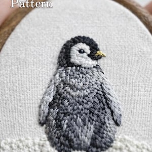 May include: An embroidery hoop containing a hand-stitched penguin chick on light-coloured fabric. The penguin features shades of black, grey, and white thread with a small yellow beak. The text PDF Pattern and lumellu.etsy.com are visible.