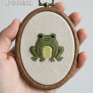 Op de afbeelding: Een ovale borduurring met een houtnerf frame en een crèmekleurig stoffen paneel. De stof bevat een met de hand geborduurde groene kikker met een lichtgele buik en zwarte ogen. De tekst "PDF Pattern" en "lumellu.etsy.com" is zichtbaar.