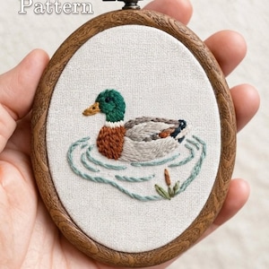 Könnte beinhalten: Ein ovaler Holzstickrahmen mit einem Stoffeinsatz, der eine handgestickte Stockente auf dem Wasser zeigt. Die Ente hat einen grünen Kopf, eine braune Brust und graue Flügel. Der Text PDF Pattern und lumellu.etsy.com ist auf dem Bild zu sehen.
