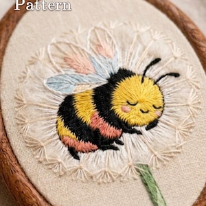Pode incluir: Um projeto de bordado num bastidor de madeira com um abelhão de riscas amarelas e pretas e asas azul-claras a descansar sobre uma flor de dente-de-leão branca. O texto PDF Pattern é visível no canto superior esquerdo do tecido.