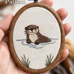 May include: An oval embroidery hoop containing a fabric panel with a hand-stitched brown otter swimming in blue water, surrounded by green reeds. The frame is dark wood with a metal hanging loop. Text reads PDF Pattern and Lumellu.