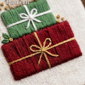 May include: An embroidery design featuring three stacked gift boxes on a light fabric background. The top box is beige, the middle is green with a white ribbon, and the bottom is red with a gold ribbon. Text reads PDF Pattern and Lumellu.