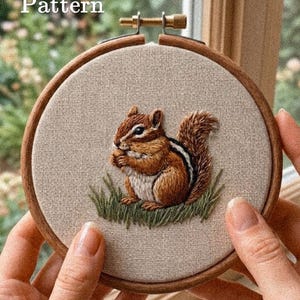 Puede incluir: Un bastidor de bordado circular de madera que contiene un panel de tela beige con un diseño bordado a mano de una ardilla listada marrón sentada en hierba verde. El texto "PDF Pattern" está impreso en blanco en la esquina superior izquierda.