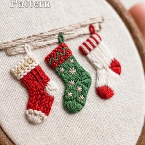 Op de afbeelding: Een borduurring met een PDF Pattern van drie hangende kerstsokken op gebroken wit stof. De sokken zijn rood met een crèmekleurige boord, groen met een rode boord en sterdetails, en wit met rode strepen en accenten. Lumellu.