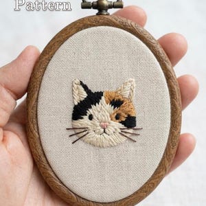 Pode incluir: Um bastidor de bordado oval com acabamento em grão de madeira contendo um rosto de gato calico bordado em tecido creme. O gato tem pelo preto, laranja e branco, um nariz rosa e bigodes castanhos. O texto PDF Pattern e Lumellu é visível.