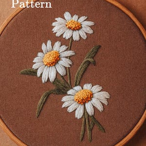 May include: A circular wooden embroidery hoop containing brown fabric with a hand-stitched design of three white daisies with yellow centers and green stems. The text Pdf Pattern is printed in white on the upper left side of the fabric.