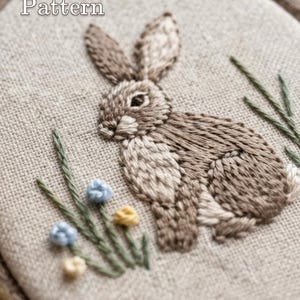 May include: An embroidery hoop containing a beige linen fabric with a hand-stitched brown rabbit, green grass blades, and small blue and yellow flowers. The text 'PDF Pattern' is at the top and 'lumellu.etsy.com' is at the bottom of the image.