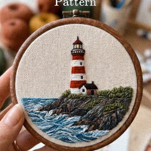 May include: A circular wooden embroidery hoop containing a fabric panel with a lighthouse design. The lighthouse has red and white horizontal stripes. Below it are dark rocky cliffs, green vegetation, and blue ocean waves. Text reads PDF Pattern.