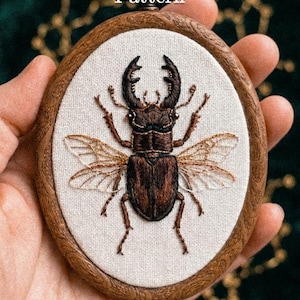 Puede incluir: Aro de bordado ovalado de madera con un diseño detallado de escarabajo ciervo marrón. El escarabajo está bordado sobre tela blanca, con intrincados detalles de alas y cuerpo. El texto "Pdf Pattern" está en la parte superior y "lumellu.com" en la inferior.