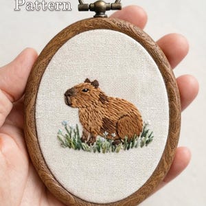 Peut inclure: Un tambour à broder ovale avec un cadre imitation bois contenant un panneau en tissu avec un capybara marron brodé dans de l'herbe verte. Le texte PDF Pattern est en haut à gauche et le texte Lumellu est en bas au centre.