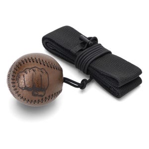 Könnte beinhalten: Ein brauner Leder-Baseball mit schwarzen Nähten und einer schwarzen Faustgrafik auf der Oberfläche. Der Baseball ist mit einer dünnen schwarzen Kordel an einem gefalteten schwarzen elastischen Stirnband mit einem kleinen schwarzen Kunststoffclip befestigt.