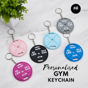 Könnte beinhalten: Eine Sammlung von sechs runden Hantelscheiben-Schlüsselanhängern in Pink, Schwarz, Hellblau, Grau und Blau. Jeder Anhänger zeigt den eingeprägten Text BUMI, 45 LBS, 20.4 KGS und GYM und ist an einem silbernen Metallring mit Kette befestigt.