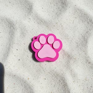 Op de afbeelding: Een felroze label in de vorm van een pootafdruk met een lichtere roze centrale voetzool en vier kleinere teenzooltjes. Het object heeft een klein rond oogje in de linkerbovenhoek voor bevestiging en een textuur op het gehele oppervlak.