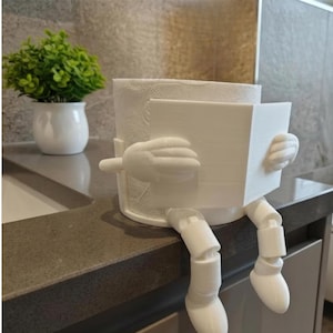 May include: A white, 3D-printed toilet paper holder shaped like a person reading a book. The holder has arms, legs, and a book-shaped front. A small, white potted plant sits in the background on a gray countertop.