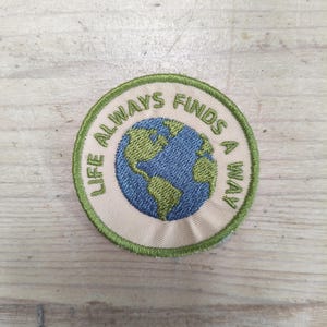 Op de afbeelding: Een ronde geborduurde patch met een beige achtergrond en een groene gestikte rand. Het midden toont een afbeelding van een blauwe en groene wereldbol, omringd door de tekst "LIFE ALWAYS FINDS A WAY" geborduurd in groen garen.