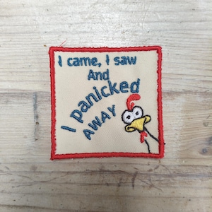 Pode incluir: Um emblema bordado quadrado com fundo bege e borda vermelha. Apresenta o texto azul "I came, I saw And I Panicked AWAY" e um gráfico bordado de uma galinha branca com bico amarelo e crista vermelha que parece surpresa.