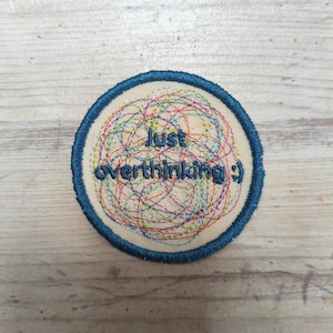 May include: A round embroidered patch with a blue border and the words "Just overthinking :)" in a light beige color. The center features a colorful, tangled design of threads in various colors.