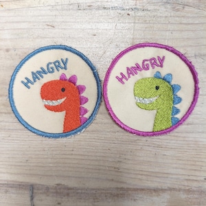 Puede incluir: Dos parches redondos bordados con un diseño de dinosaurio. Uno tiene un dinosaurio rojo con borde azul y la palabra "HANGRY" en azul. El otro tiene un dinosaurio verde con borde rosa y la palabra "HANGRY" en rosa.