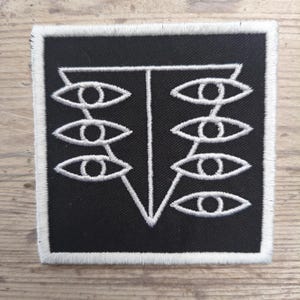 Könnte beinhalten: Ein quadratischer schwarzer Aufnäher mit einem weißen gestickten Design. Das Design zeigt eine zentrale vertikale Linie mit einem Dreieck am unteren Ende und einer horizontalen Linie am oberen Ende. Sechs stilisierte Augen sind auf beiden Seiten der vertikalen Linie angeordnet. Der Aufnäher hat einen weißen Rand.