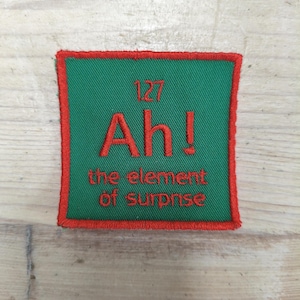 May include: A green square patch with a red border. The patch features the text "Ah! the element of surprise" in red, with the number "127" above. The patch is likely an embroidered item.