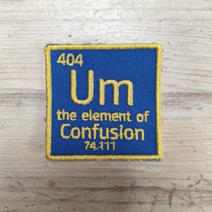May include: A blue square patch with gold trim and embroidered text. The text reads "404 Um the element of Confusion 74.111". The patch is likely for a jacket or bag.