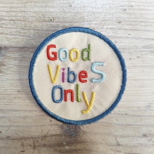 Pode incluir: Um emblema bordado circular com fundo creme e uma borda costurada em azul escuro. O texto "Good VibeS Only" está bordado no centro usando fios de várias cores, incluindo vermelho, amarelo, azul, verde e azul claro.