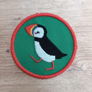 Op de afbeelding: Geborduurde patch met een cartoonpapegaaiduiker. De vogel is zwart, wit en lichtblauw, met een rode, gele en blauwe snavel. De patch heeft een groene achtergrond met een rode rand.