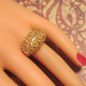 May include: A gold-colored ring with a cluster of small, round embellishments. The ring is oval-shaped and sits on a person's finger. The background is a patterned design with shades of brown and red.