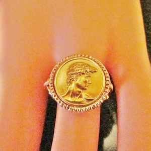 May include: A gold-colored ring featuring a detailed profile of a person on a circular medallion. The ring has a textured band and a decorative border around the medallion. The ring is a classic piece of jewelry.