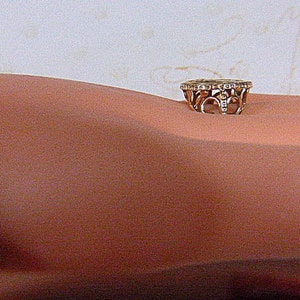 May include: A gold-toned ring with a decorative design and small, clear stones. The ring is set on a skin-toned surface. The ring's design features openwork patterns and a band of small stones around the top.