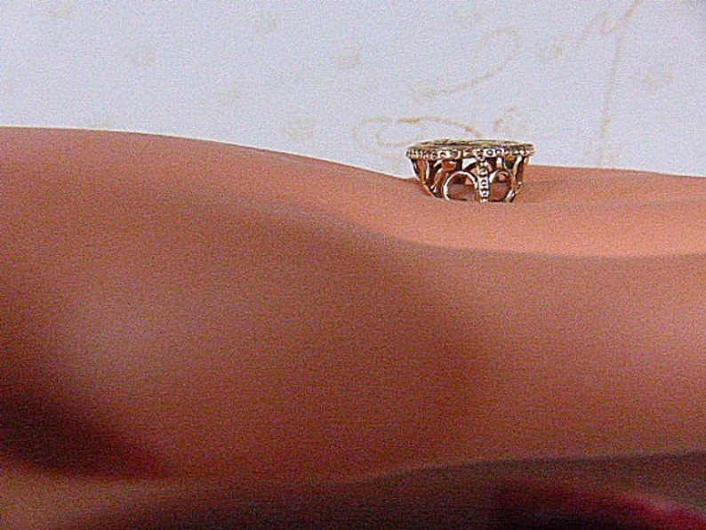 May include: A gold-toned ring with a decorative design and small, clear stones. The ring is set on a skin-toned surface. The ring's design features openwork patterns and a band of small stones around the top.