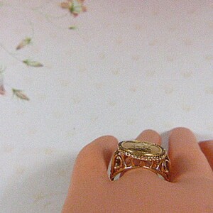 May include: A gold-toned ring featuring a coin-like centerpiece. The ring has an ornate, openwork design around the coin, with a detailed pattern. The ring is shown on a person's finger, with a soft, neutral background.