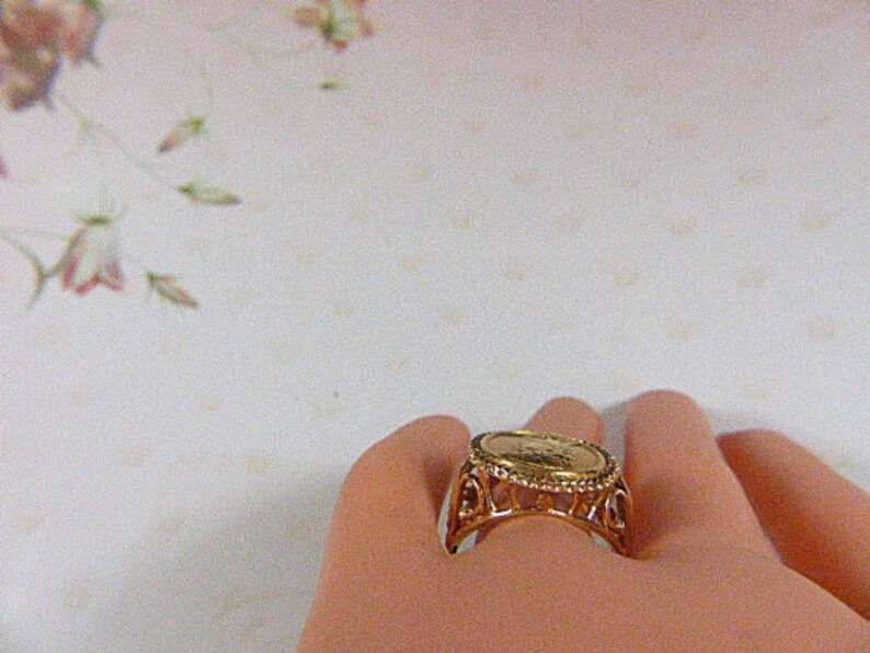 May include: A gold-toned ring featuring a coin-like centerpiece. The ring has an ornate, openwork design around the coin, with a detailed pattern. The ring is shown on a person's finger, with a soft, neutral background.
