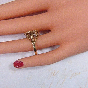 May include: A gold-colored ring with an ornate filigree design. The ring features a detailed, openwork pattern around a central stone setting. The ring is displayed on a hand with red nail polish.