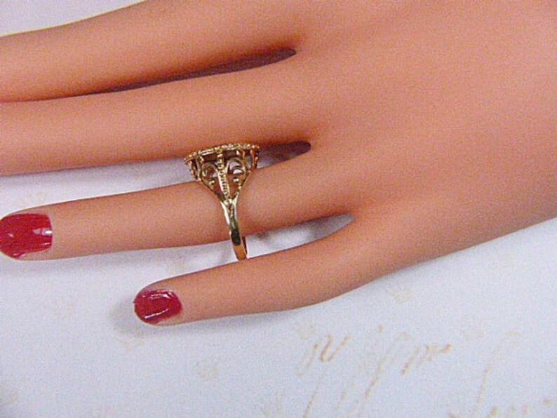 May include: A gold-colored ring with an ornate filigree design. The ring features a detailed, openwork pattern around a central stone setting. The ring is displayed on a hand with red nail polish.
