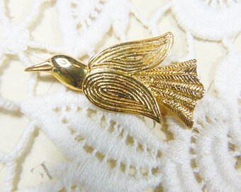 May include: A gold-toned bird brooch with intricate details. The bird is stylized with a long, thin beak and a rounded body. The wings are spread out and have a feathery texture.