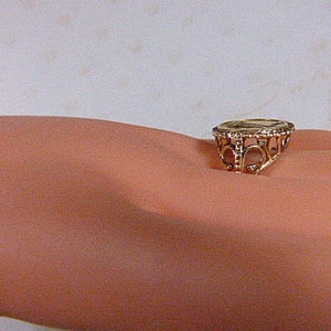 May include: A gold-colored ring with an ornate filigree design. The ring features a circular central element and decorative openwork around the band. The ring is shown on a person's skin.