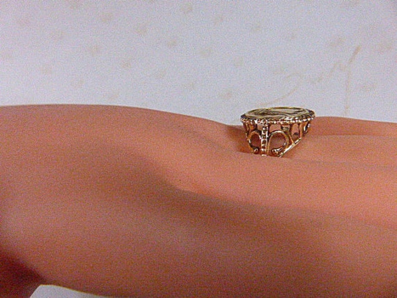 May include: A gold-colored ring with an ornate filigree design. The ring features a circular central element and decorative openwork around the band. The ring is shown on a person's skin.