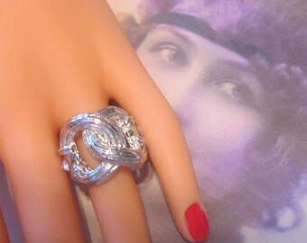 May include: A silver ring with a knot design and clear stones. The ring is on a hand wearing red nail polish.