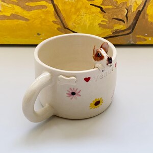 Könnte beinhalten: Cremefarbene Keramiktasse mit Henkel, verziert mit einer kleinen Hundefigur, die über den Rand schaut. Die Tasse ist mit bemalten Blumen, Herzen und dem Namen "Venus" versehen. Ein bezauberndes und einzigartiges Geschenk für Hundeliebhaber.