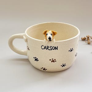Puede incluir: Taza de cerámica color crema con una figura de perro marrón y blanco en el borde. La taza presenta huellas de patas negras y el nombre "CARSON". La taza tiene un asa curva.