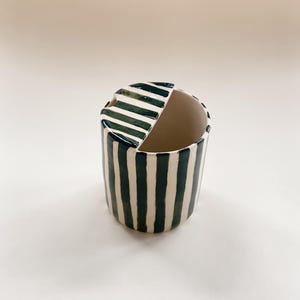 May include: A cylindrical ceramic container featuring a hand-painted pattern of vertical dark green stripes on a cream-colored background. The top of the vessel is partially covered by a matching striped lid, leaving a semi-circular opening exposed.