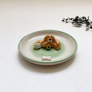 May include: A ceramic dish with a light green rim and a green and white interior. Three brown and tan dog figurines are clustered together, with a small bone-shaped figurine at the bottom. The dish is likely for decorative purposes.